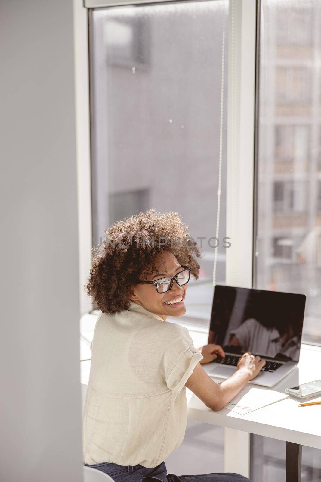 Happy Afro American lady wearing glasses and sitting at work desk while typing on laptop in the office