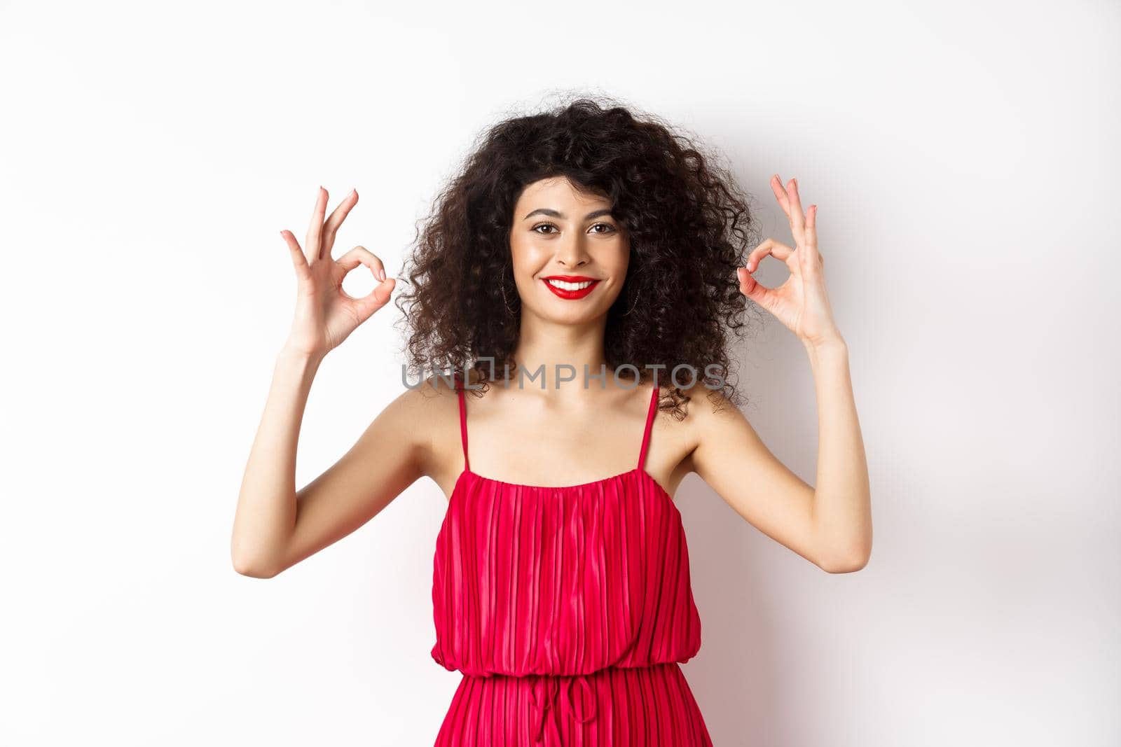 Beautiful woman in red dress, smiling and showing okay gestures, like something good, praising good job, standing over white background by Benzoix