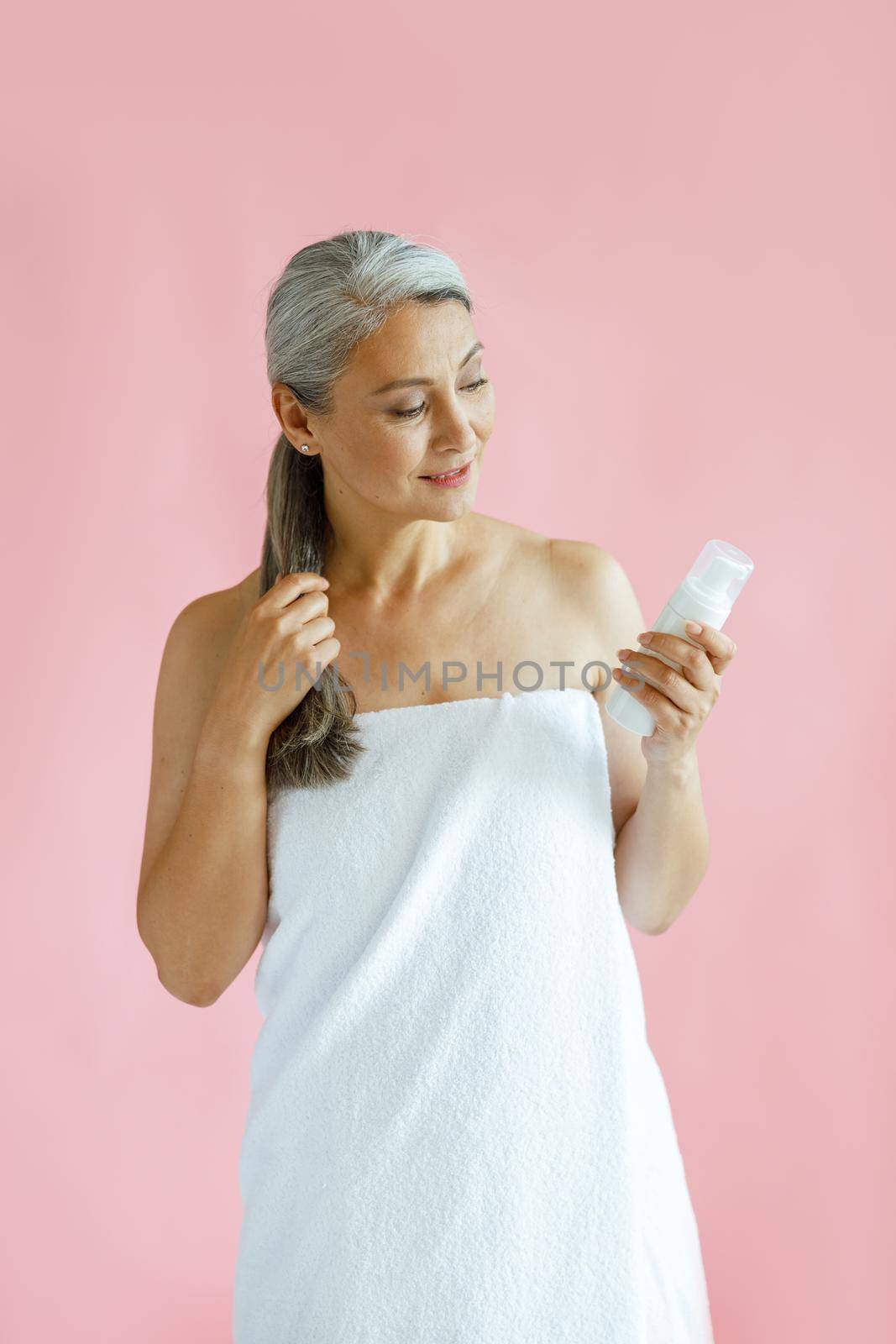 Cheerful silver haired middle aged Asian woman wrapped with terry towel looks at body care product on pink background in studio. Mature beauty lifestyle