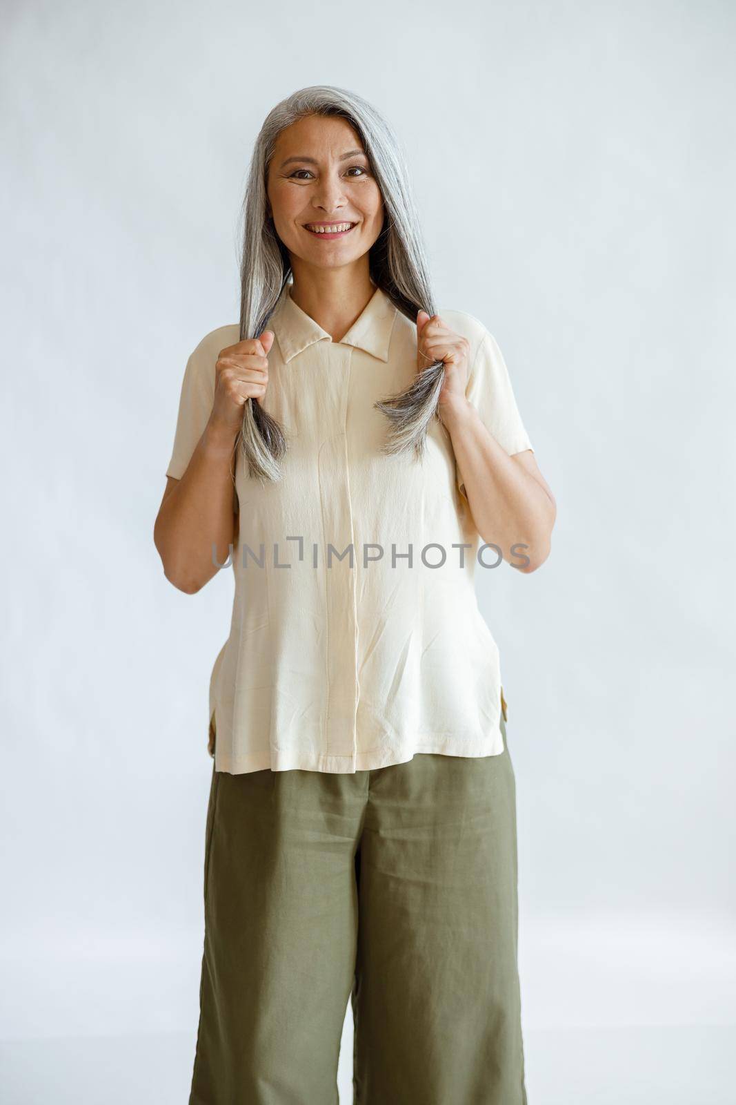 Smiling middle aged Asian woman in casual clothes holds long healthy grey hair standing on light background in studio. Mature beauty lifestyle