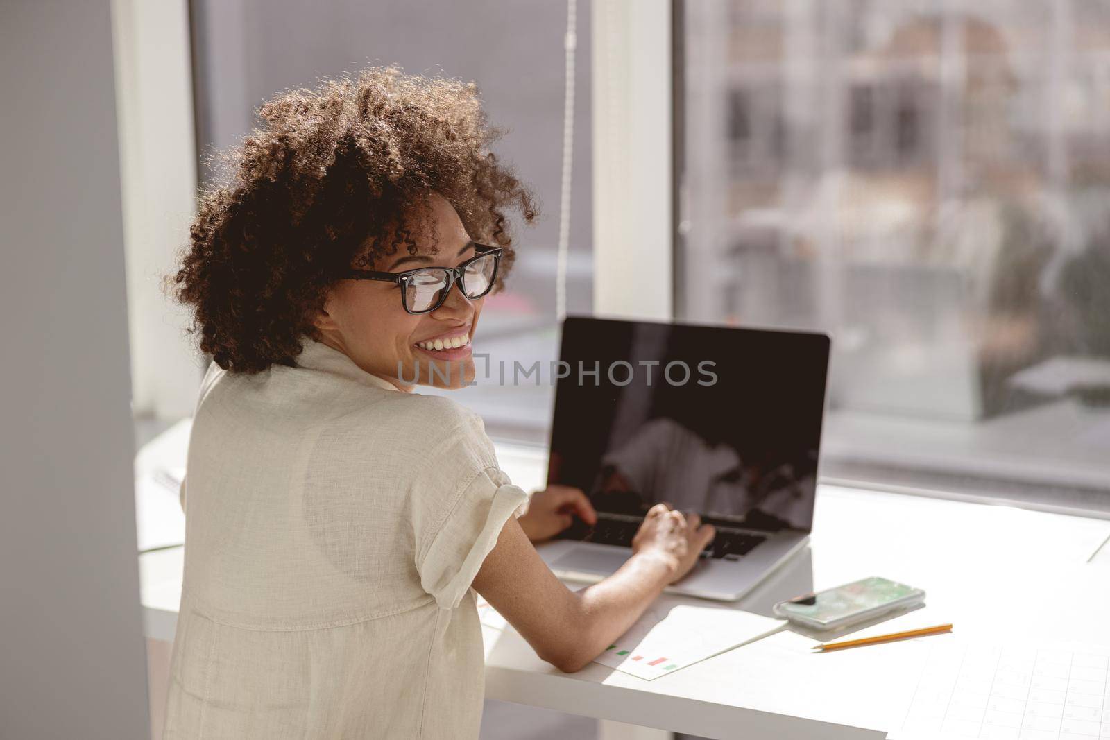 Happy Afro American lady in glasses typing on laptop while sitting at her workplace. Copy space