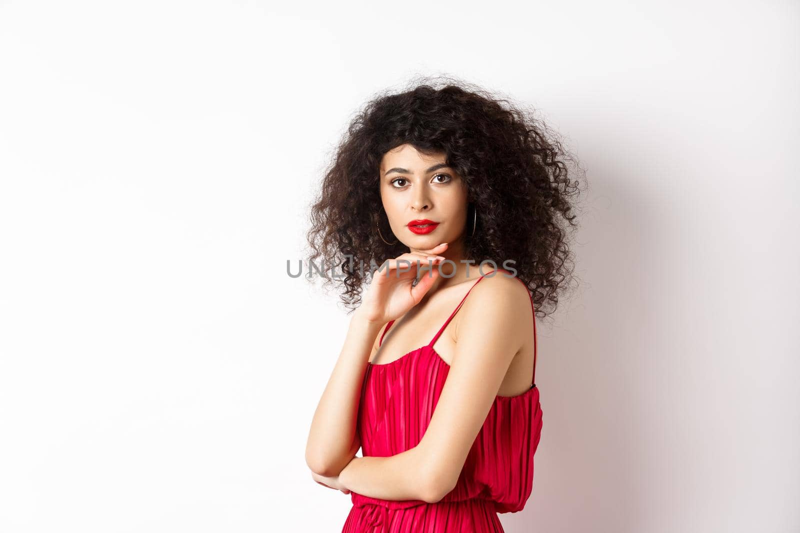 Beauty and fashion. Elegant lady with curly hair and red lips, fashionable makeup, wearing dress, gently touching chin and looking sensual at camera, white background by Benzoix