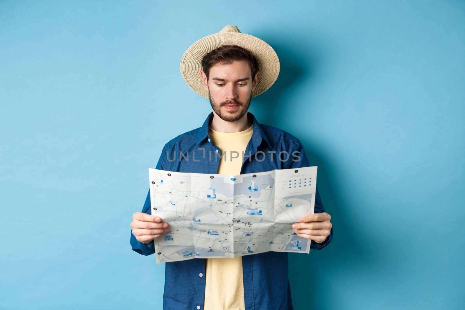 Handsome tourist with summer hat reading map, looking for sightseeing on vacation, standing on blue background.