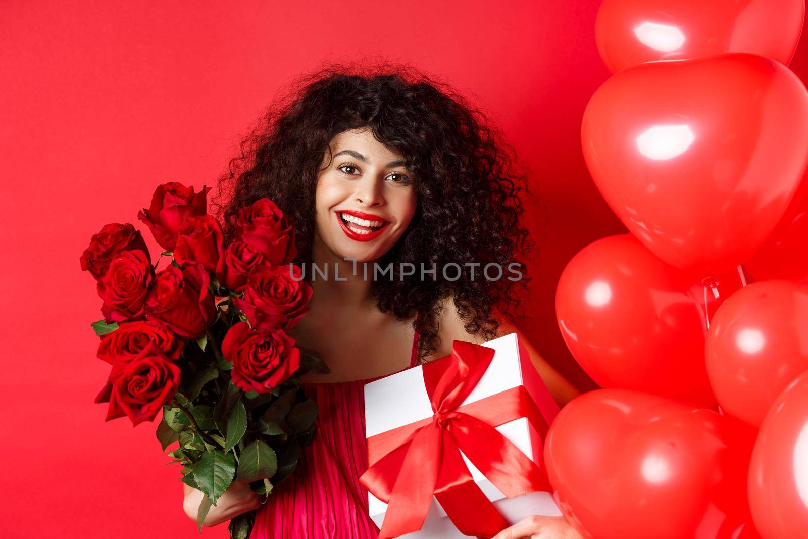 Happy Valentines day. Romantic girl with presents from lover, holding bouquet of roses and gift box, standing near cute red hearts on studio background.