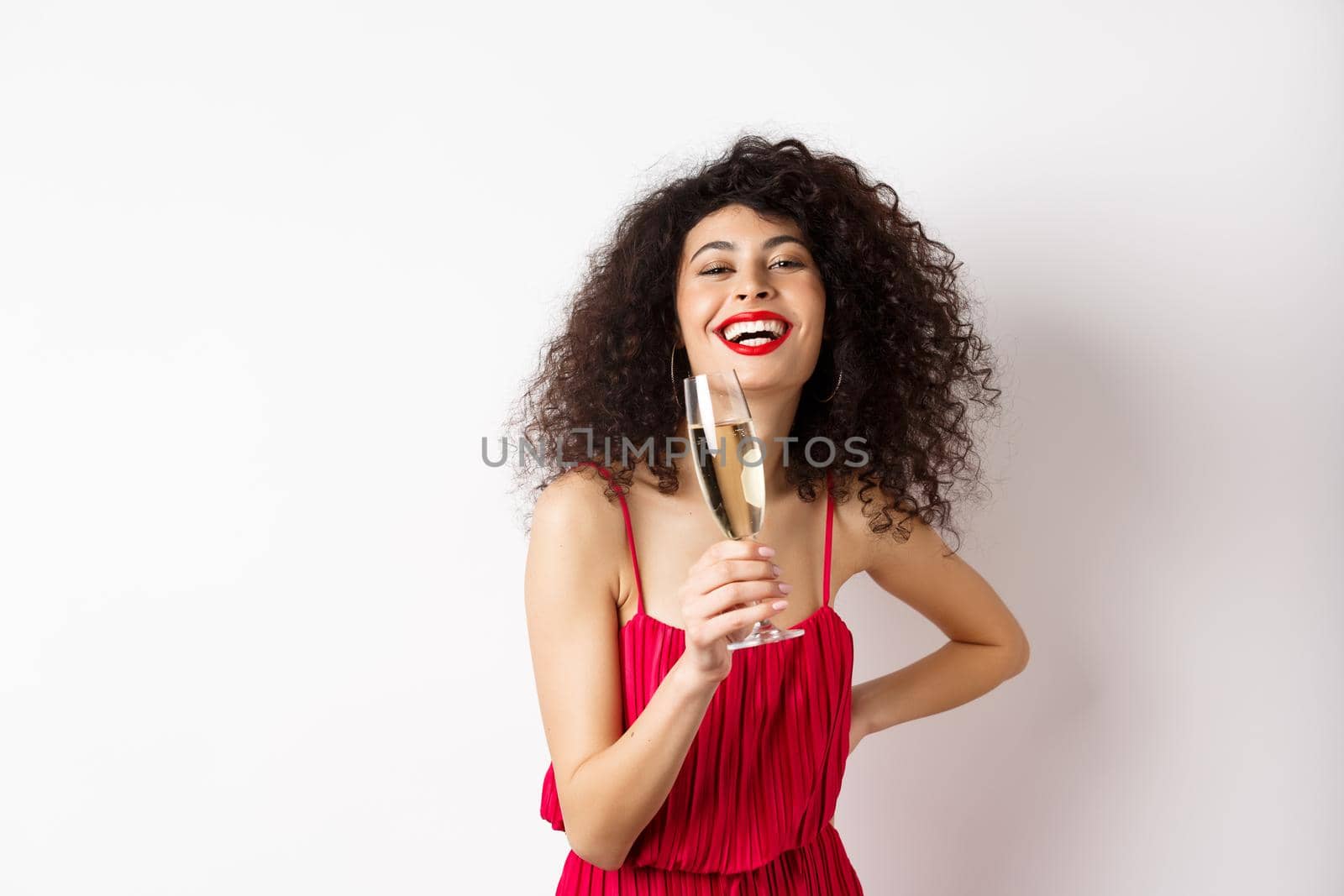 Cheerful elegant woman with dark curly hair, wearing party dress, celebrating anniversary on valentines day, drinking champagne from glass and smiling at camera, white background.