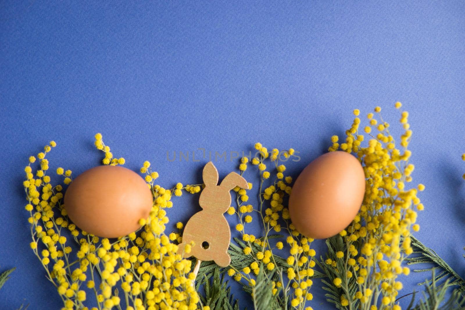 Easter background, eggs and a wooden hare on a blue background, decorated with Mimosa flowers, flatlay, top view, empty space for text. happy Easter.