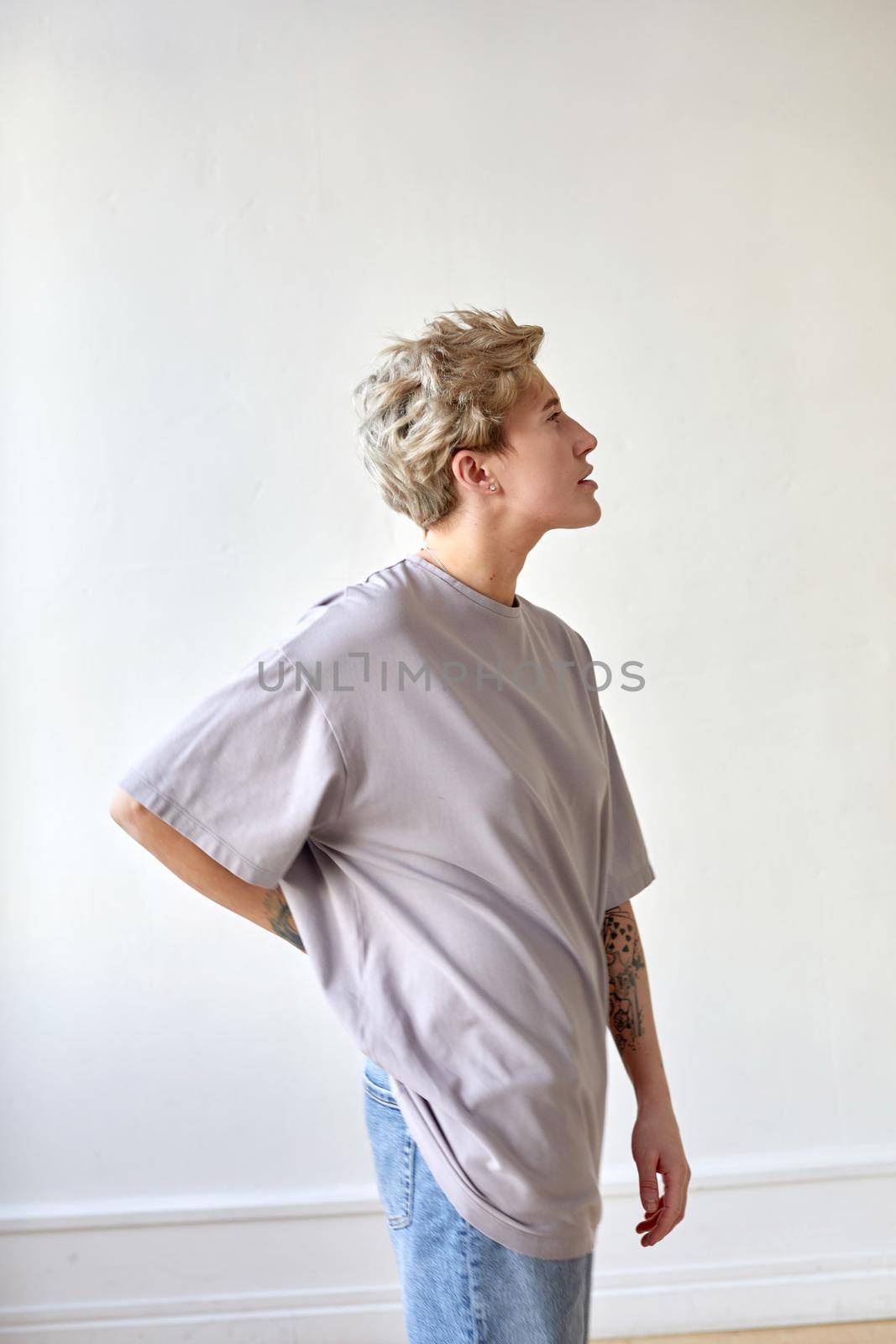 Side view of androgynous female in baggy t shirt and jeans holding hand on waist and looking away against white wall at home