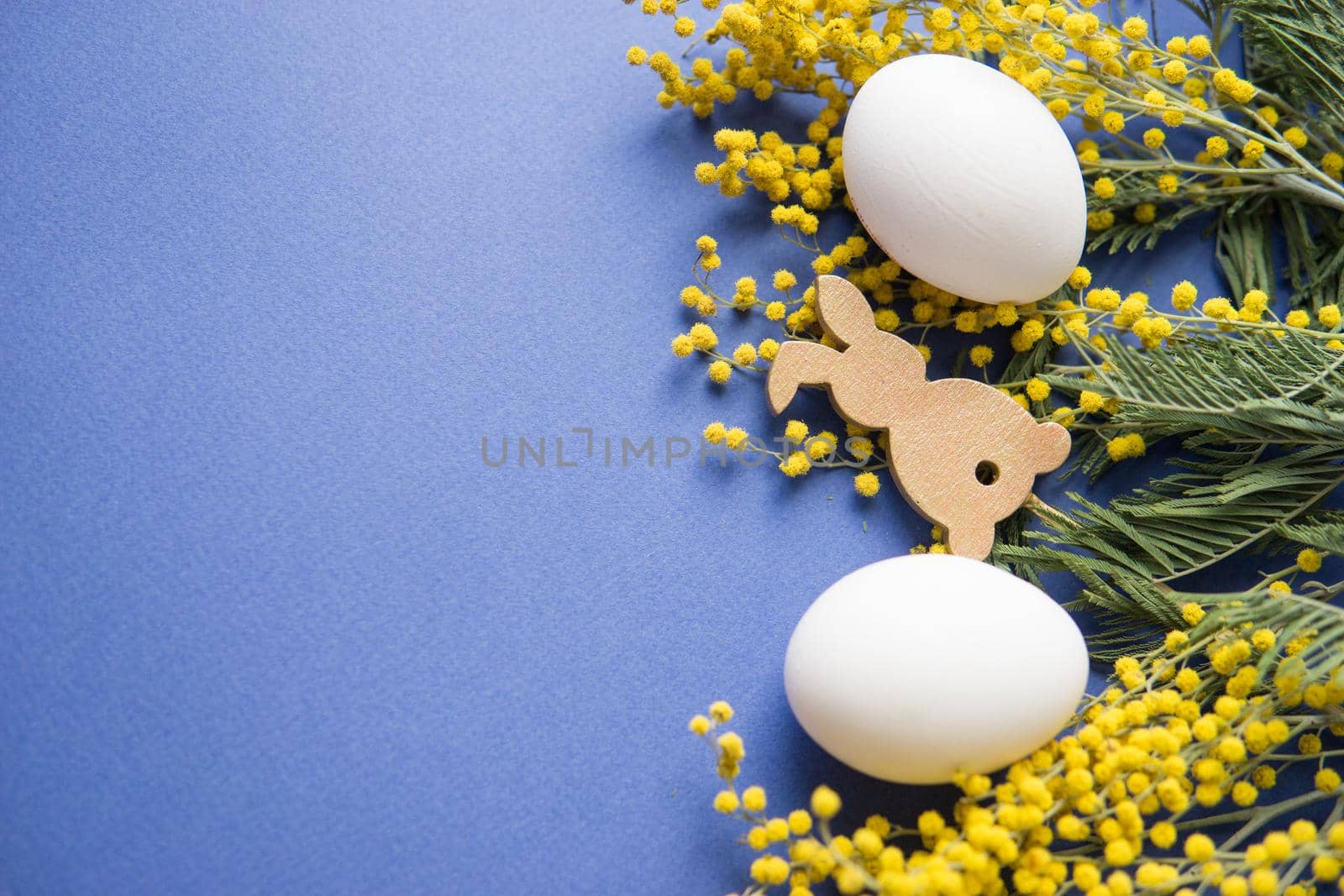 Easter background, white eggs and a wooden hare on a blue background, decorated with Mimosa flowers, flatlay, top view, empty space for text. happy Easter.