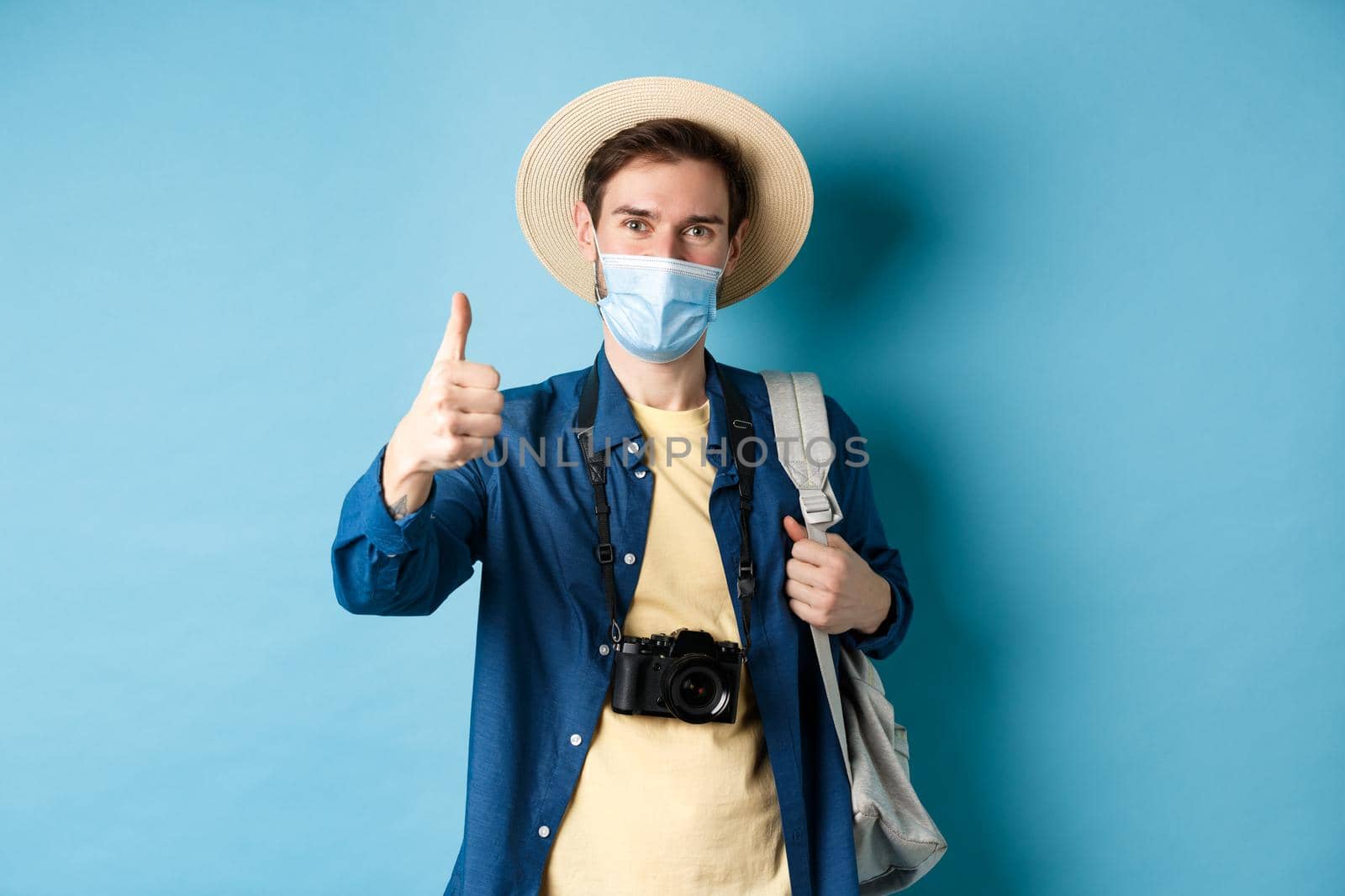 Covid-19 and summer holidays concept. Cheerful tourist in straw hat and medical mask praise and recommend good vacation hotel, showing thumb up, going on travel with backpack.