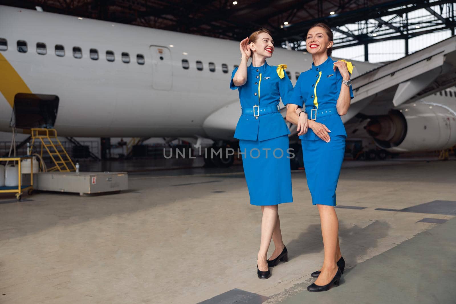 Full length shot of two air stewardesses in stylish blue uniform smiling, standing together in front of passenger aircraft in hangar at the airport. Occupation concept