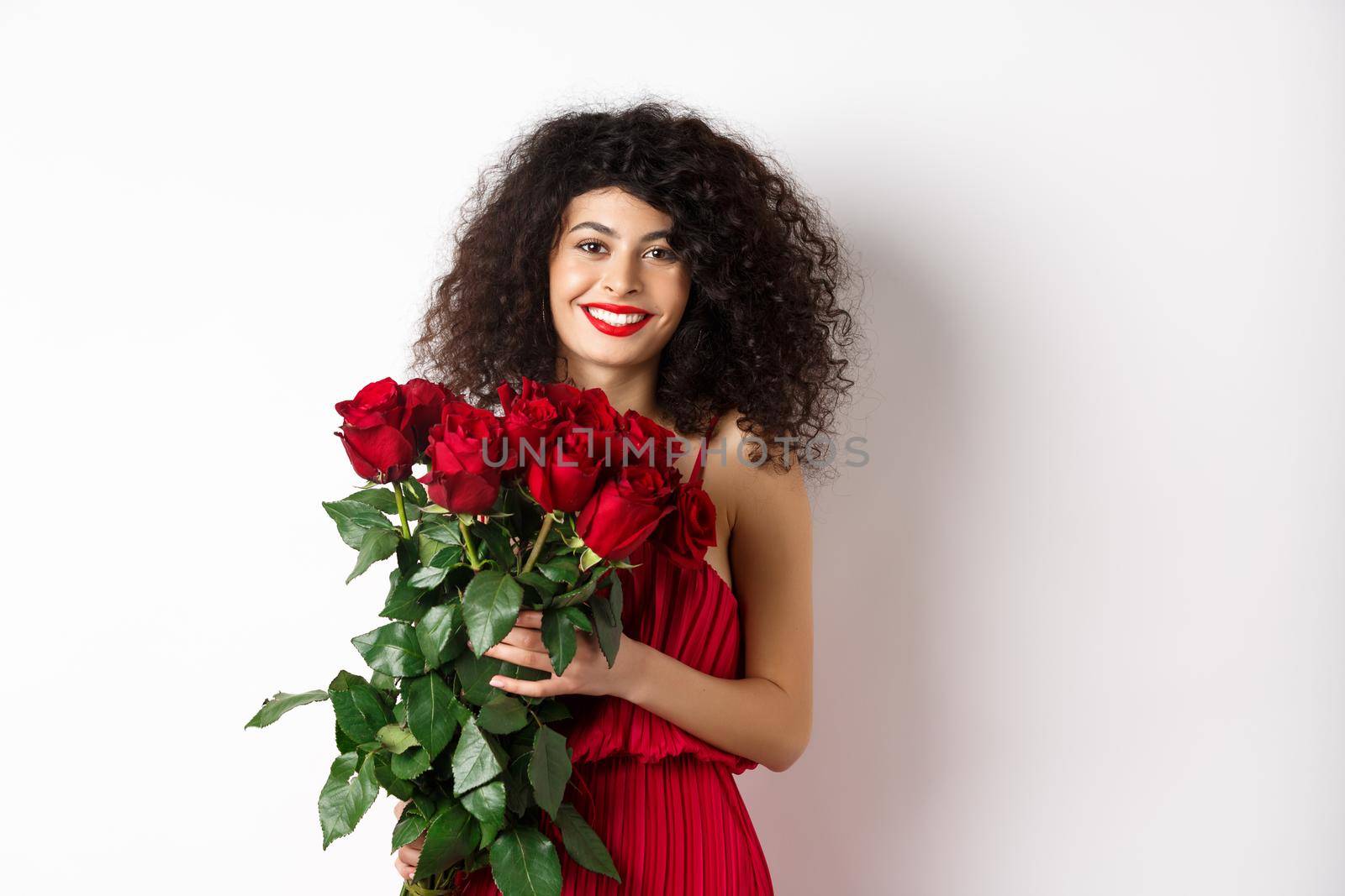 Gorgeous caucasian woman in red evening dress receiving bouquet of flowers. Girl with red roses smiling at camera, having romantic date on Valentines lover day, white background by Benzoix