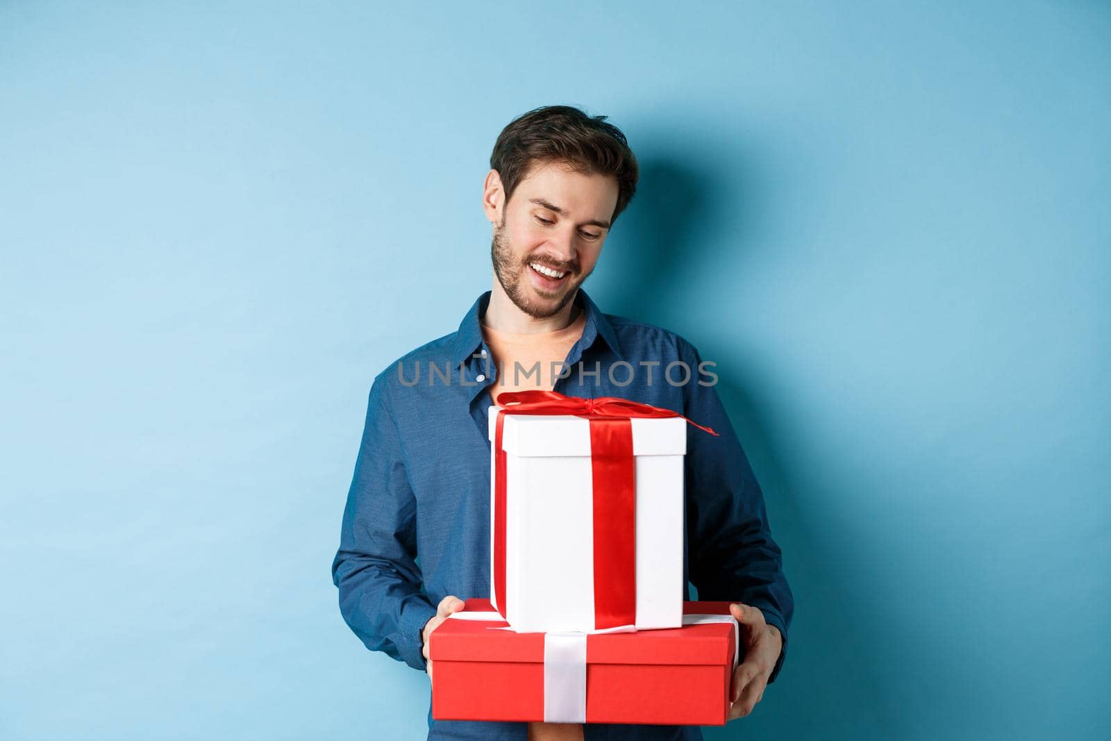 Romantic young man with beard, looking happy at gift boxes on valentines day, giving presents to lover, standing over blue background.