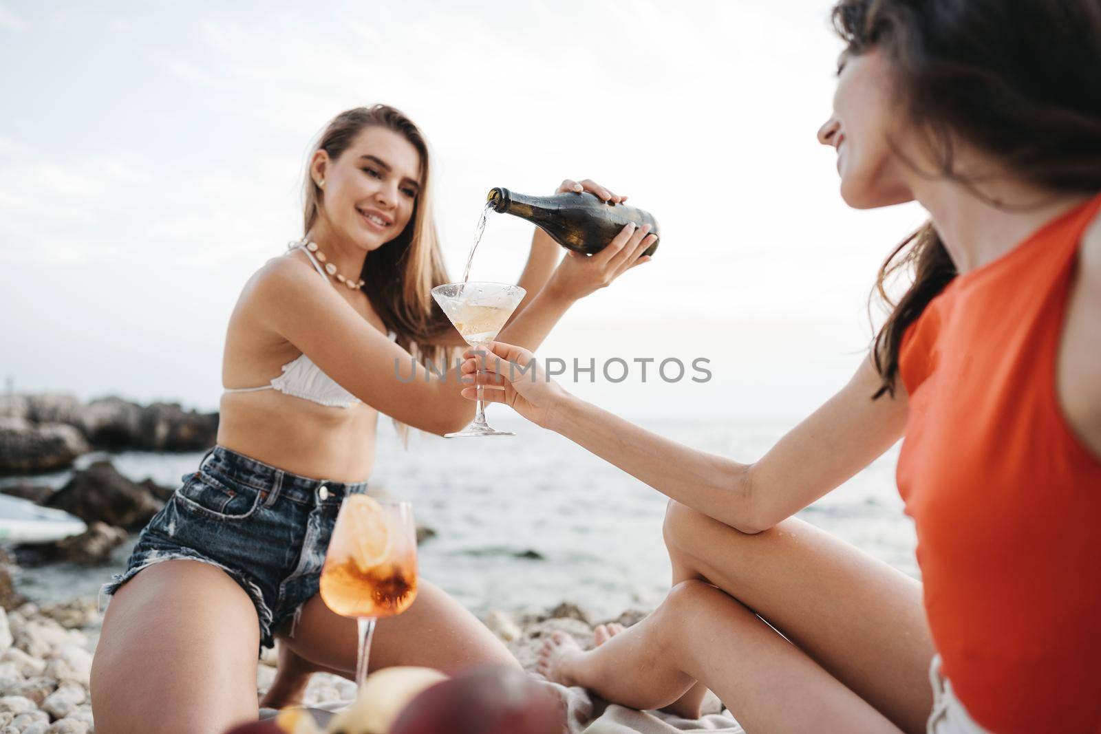 Two young female friends having picnic on a beach drinking cocktails by Fabrikasimf