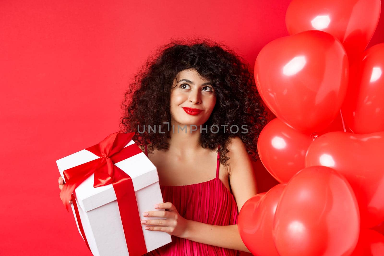 Valentines day and love concept. Beautiful woman with romantic makeup, holding gift box from secret admirer and looking at upper left corner dreamy, standing with heart balloons on red background by Benzoix