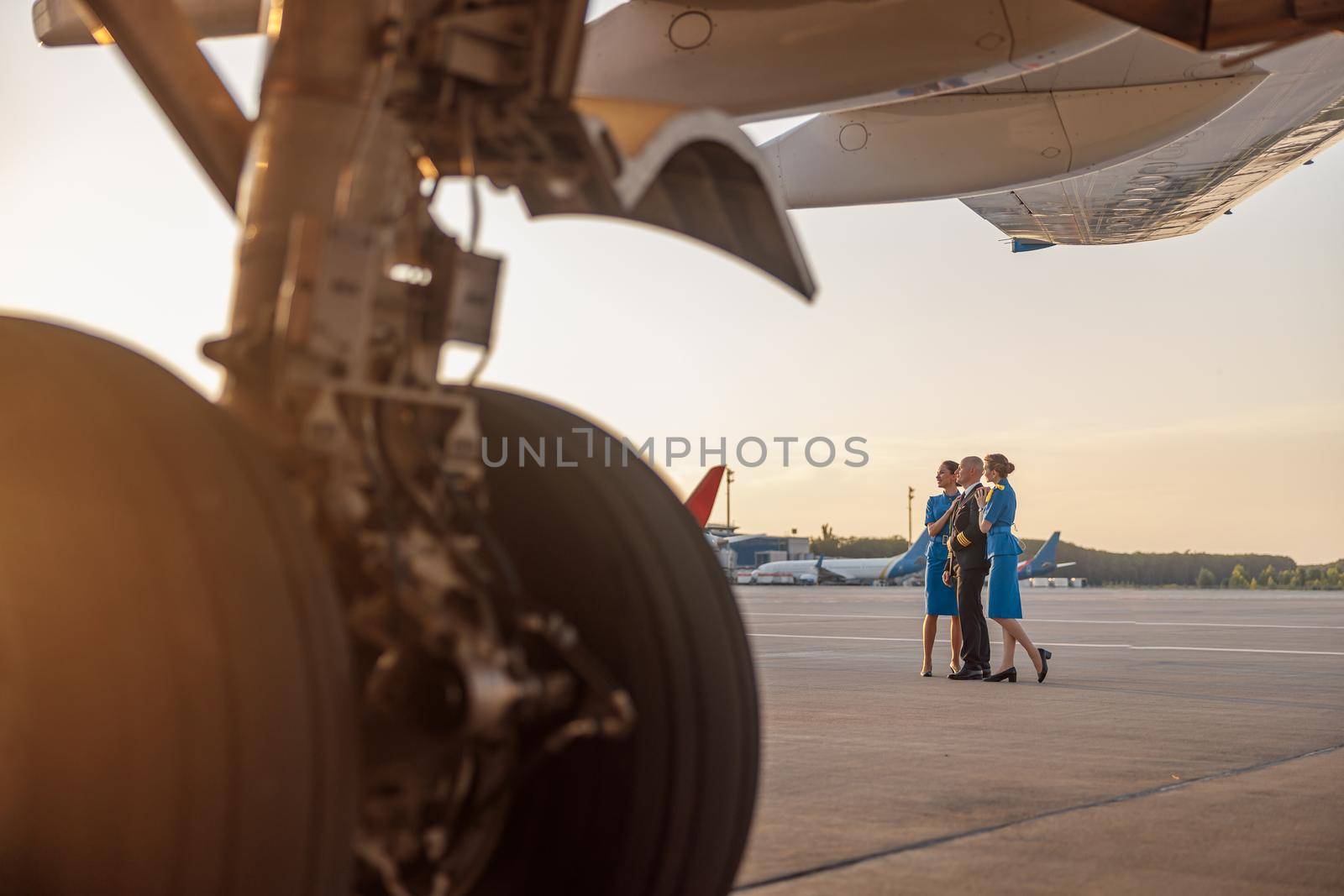 Full length shot of male pilot posing for photoshoot together with two air hostesses in blue uniform, standing in an airport terminal at sunset by Yaroslav_astakhov