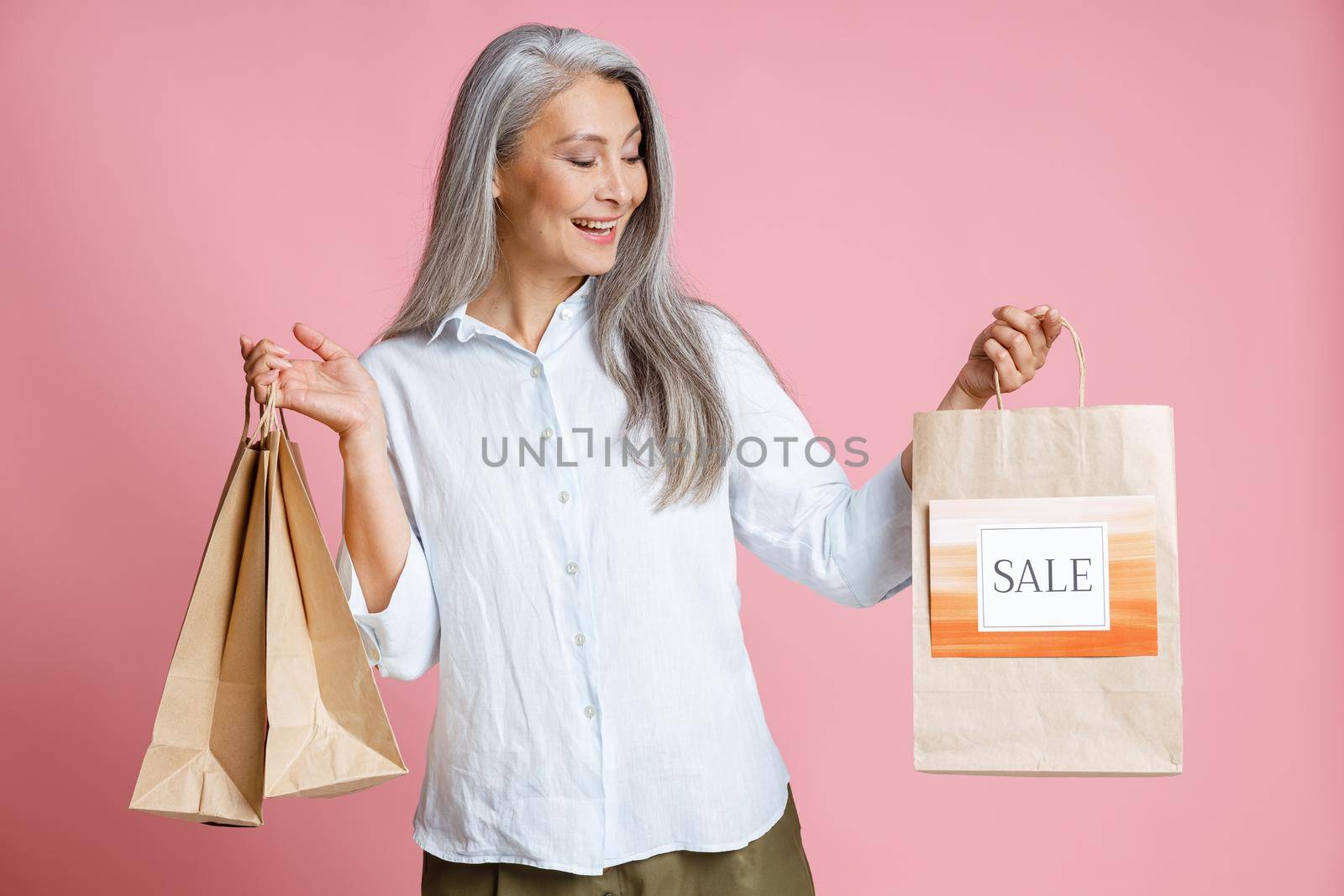 Cheerful mature Asian woman holds paper shopping bags with colorful Sale sign posing on pink background in studio