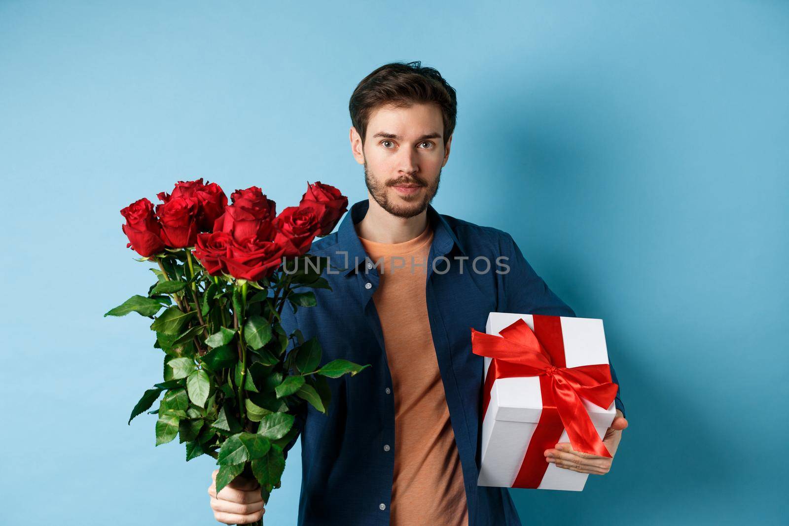 Romance and valentines day. Man presenting bouquet of red roses to lover. Boyfriend bring flowers and gift on romantic date, standing over blue background.