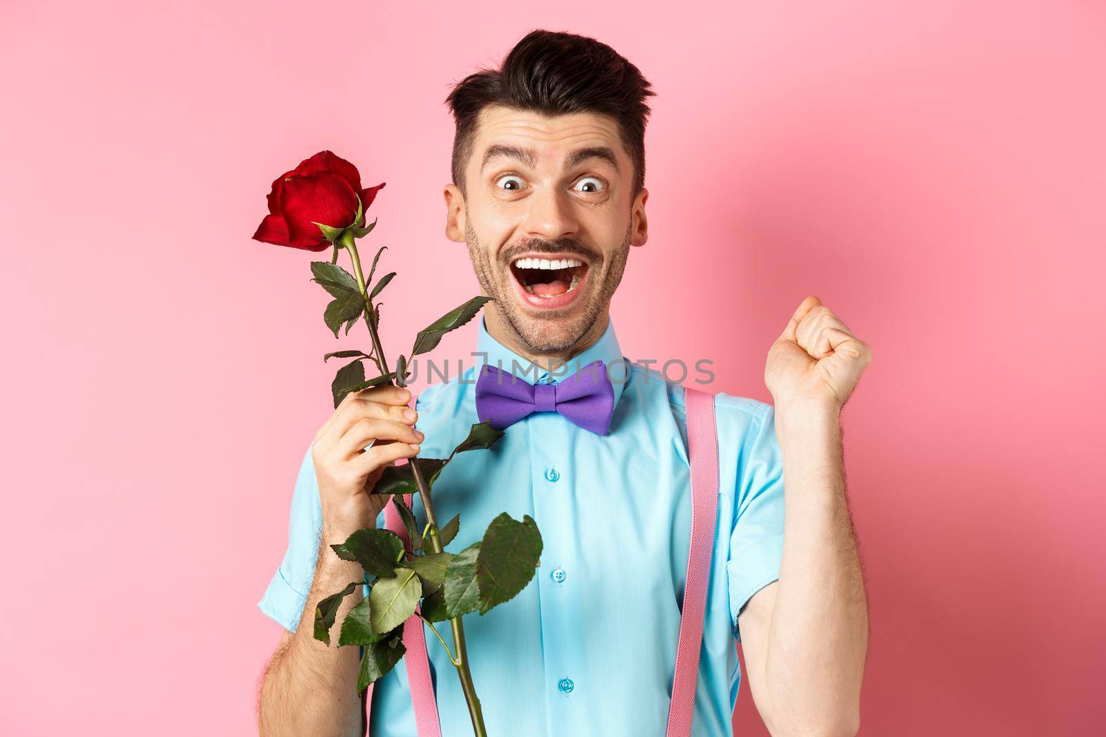 Valentines day and romance concept. Excited boyfriend jumping from happiness on romantic date, holding red rose and celebrating, standing on pink background by Benzoix