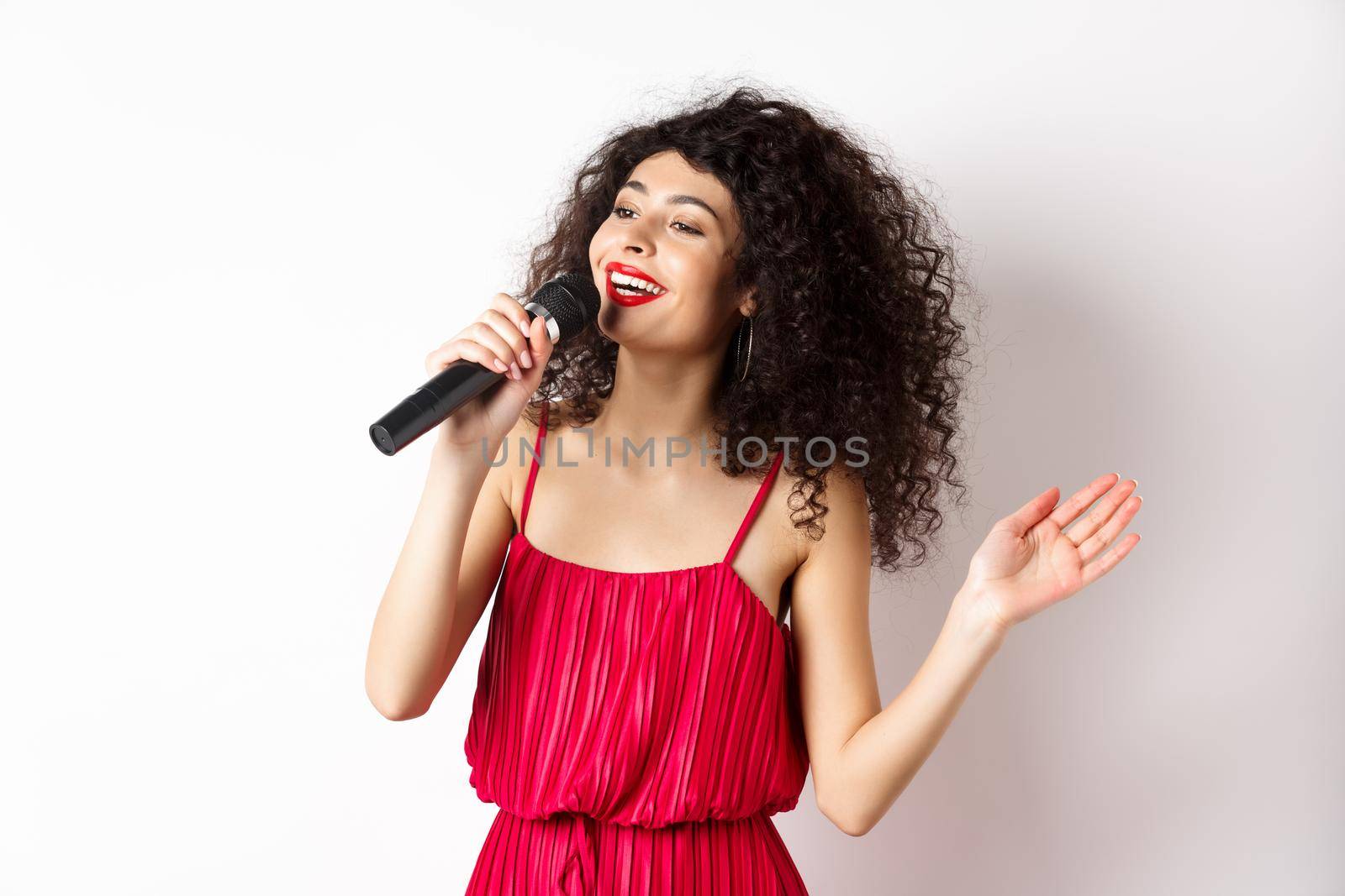 Elegant curly-haired woman in red dress singing in microphone, looking aside and smiling happy, standing on white background.