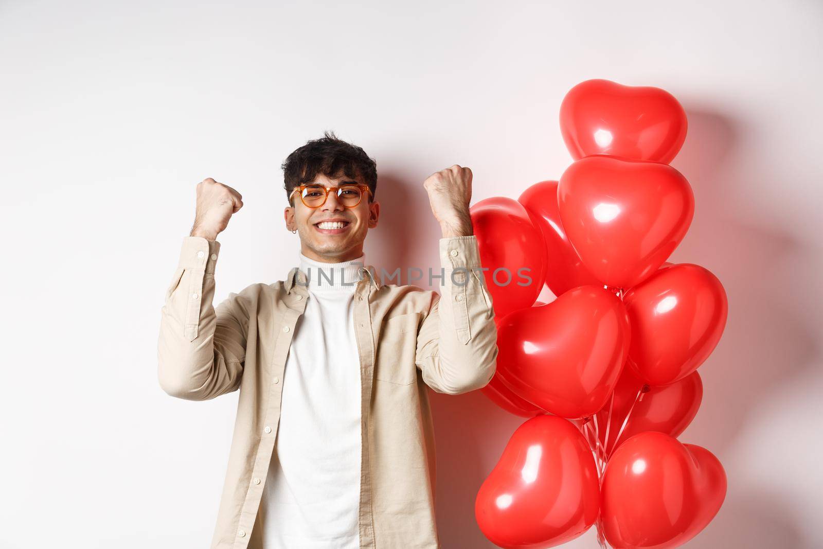Valentines day. Satisfied young man saying yes, triumphing and celebrating on lovers date, making fist pump and smiling pleased, standing near heart balloons on white background by Benzoix