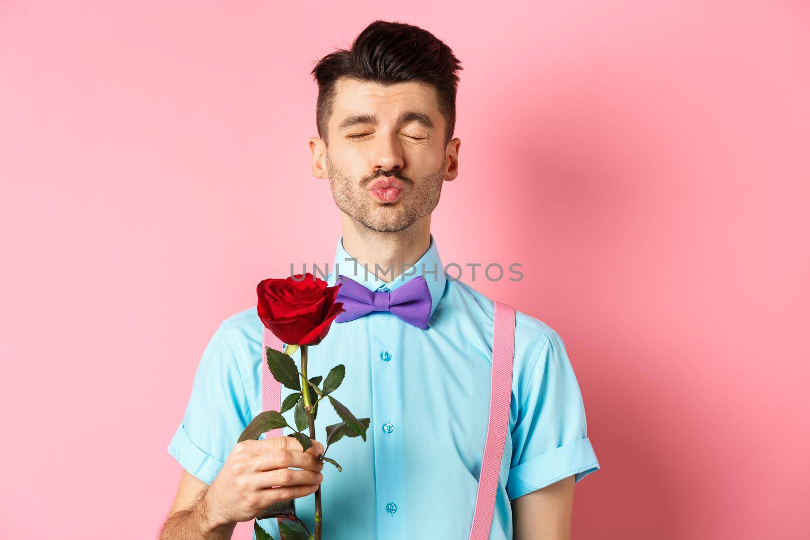 Cute and funny man waiting for kiss from lover on Valentines day, holding beautiful red rose for girlfriend, standing over pink background by Benzoix