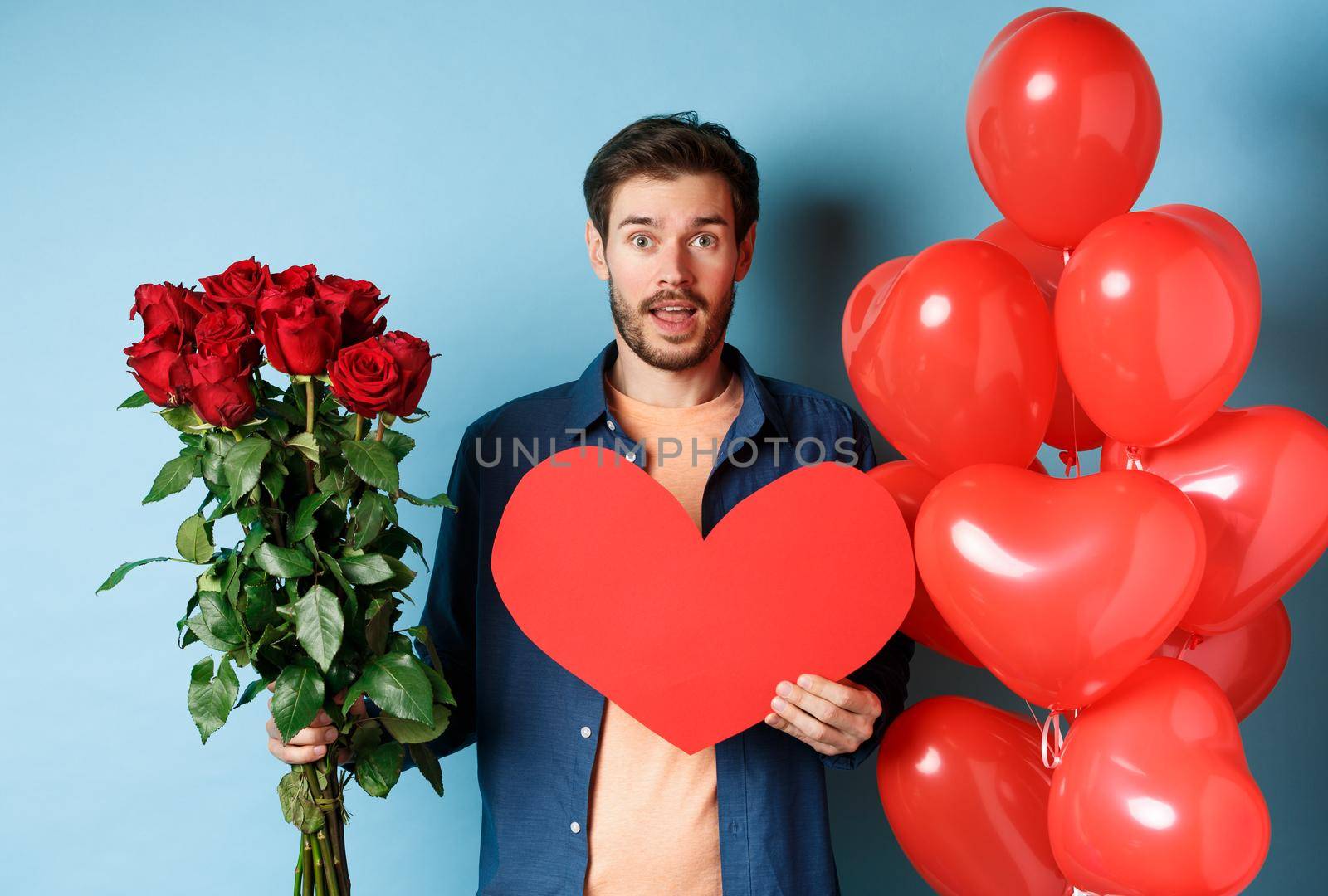 Man in love bring surprise gifts on romantic date, holding bouquet of red roses and valentine red heart, standing near balloons and looking at lover, blue background.