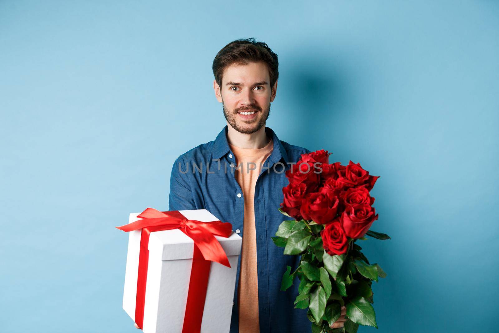 Love and Valentines day concept. Charming young man giving gift and bouquet of roses to girlfriend, standing over blue background.