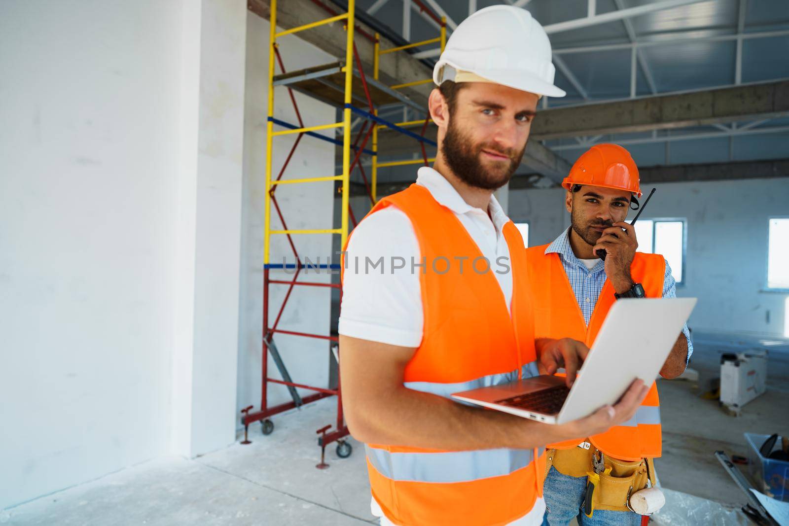 Two specialists supervisors in hardhats using laptop at construction site for work by Fabrikasimf