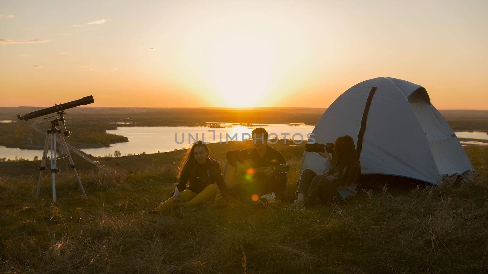 Group of friends sitting on the grass near the tent chilling out at sunset, close up