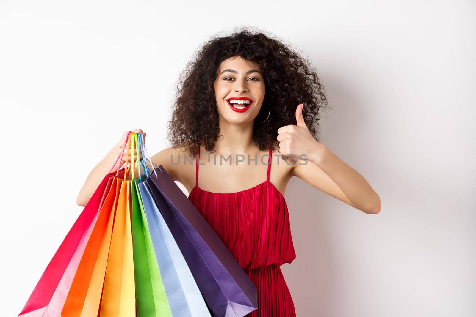 Fashionable woman in red dress going shopping, holding bags and showing thumbs up, recommend store, standing on white background by Benzoix