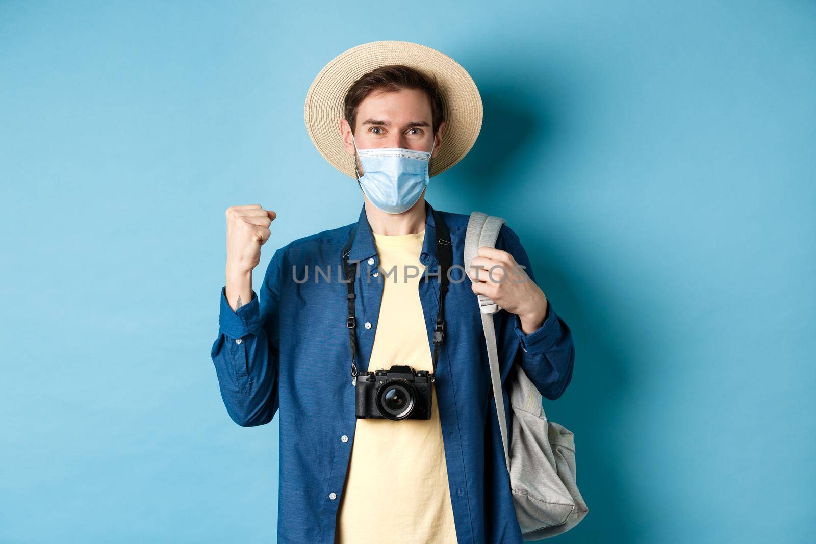 Covid-19 and summer holidays concept. Happy young man travelling abroad in medical mask, saying yes and raising fist up joyfully, cheerful about vacation, blue background by Benzoix