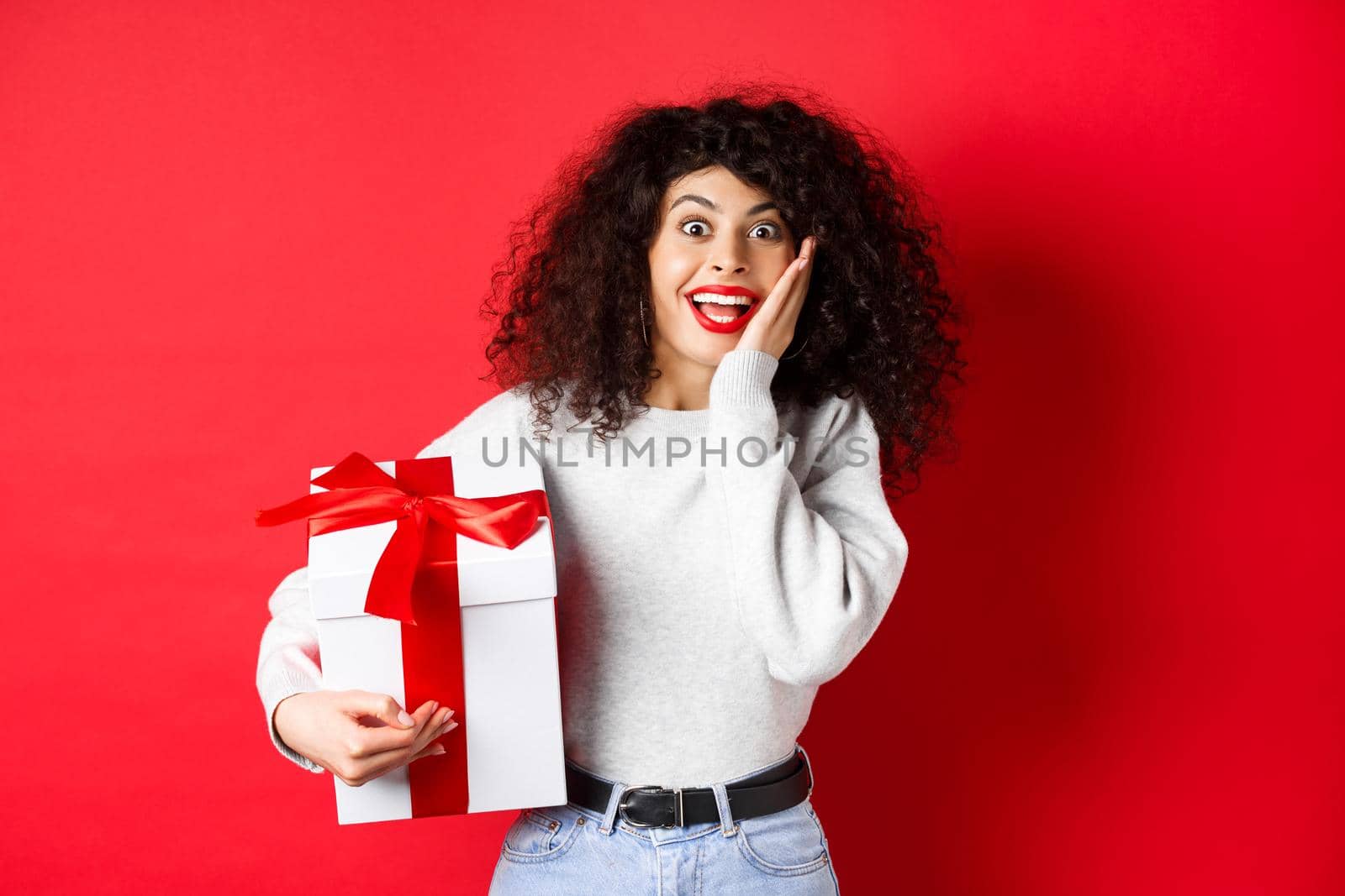 Surprised pretty girl receive Valentines day romantic gift, holding present box and looking with disbelief and happiness at camera, standing on red background by Benzoix