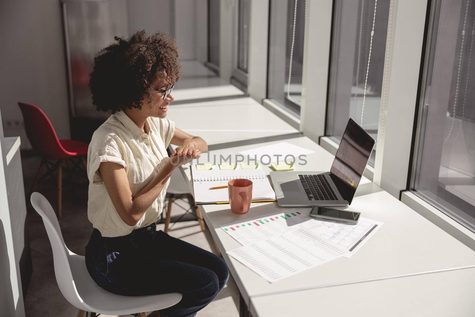 Smiling multiethnic woman learning and communicating in sign language online while sitting near window in the office