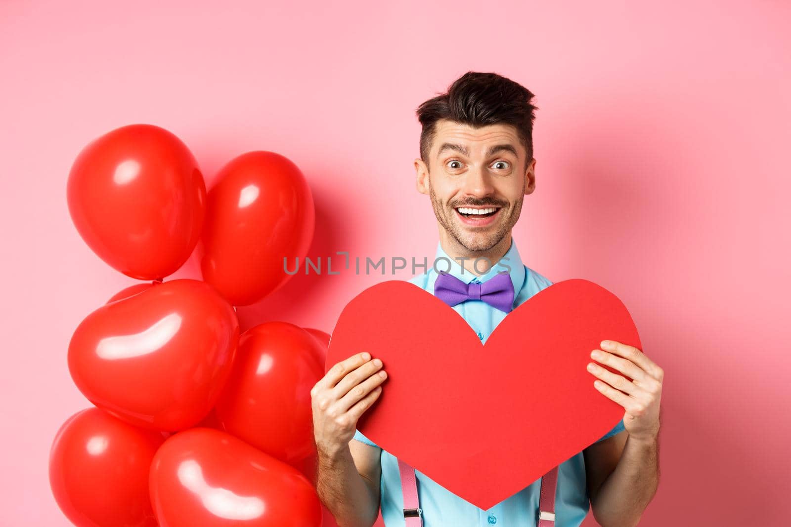 Valentines day concept. Cute young man in bow-tie showing big red heart postcard and say love you, smiling happy at camera, standing on romantic pink background.