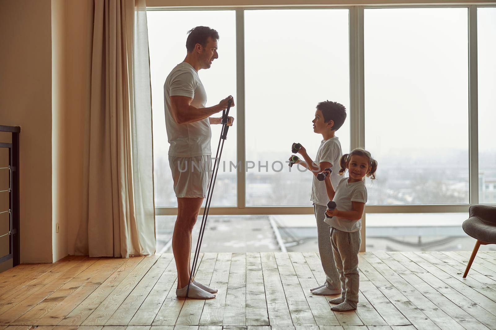 Father with resistance bands is teaching happy daughter and son exercising with dumbbells before window