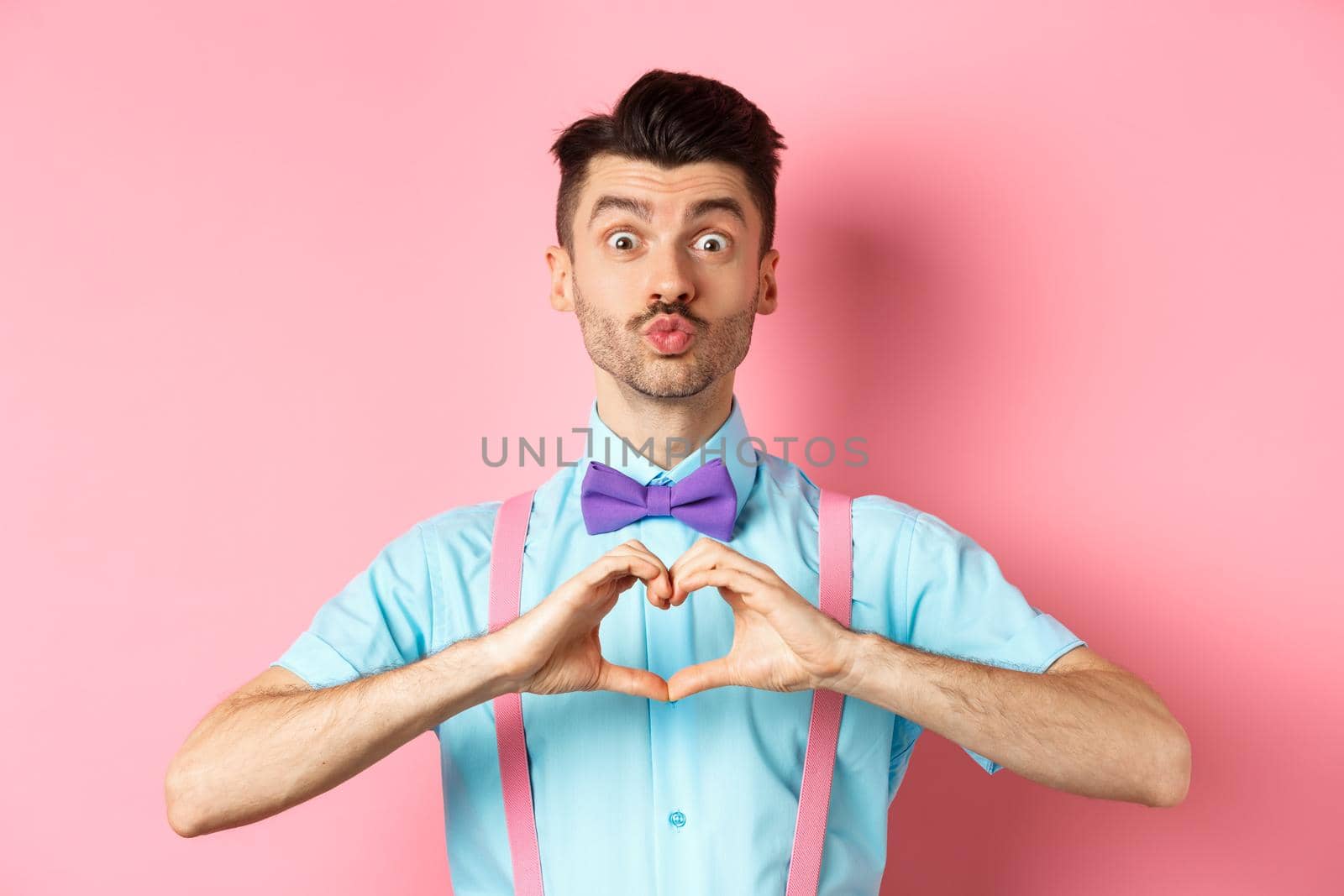 Happy Valentines day. Funny young man waiting for lovers kiss, pucker lips and show heart sign, I love you gesture, express feelings on date, pink background.