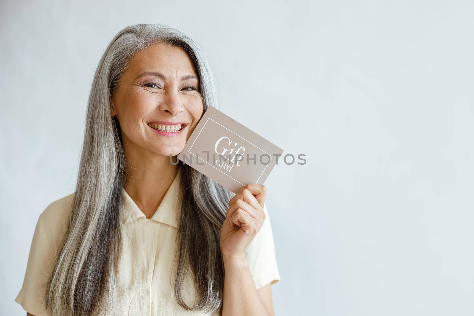Pretty smiling mature Asian lady holds gift card posing on light grey background in studio, space for text. Shopping certificate
