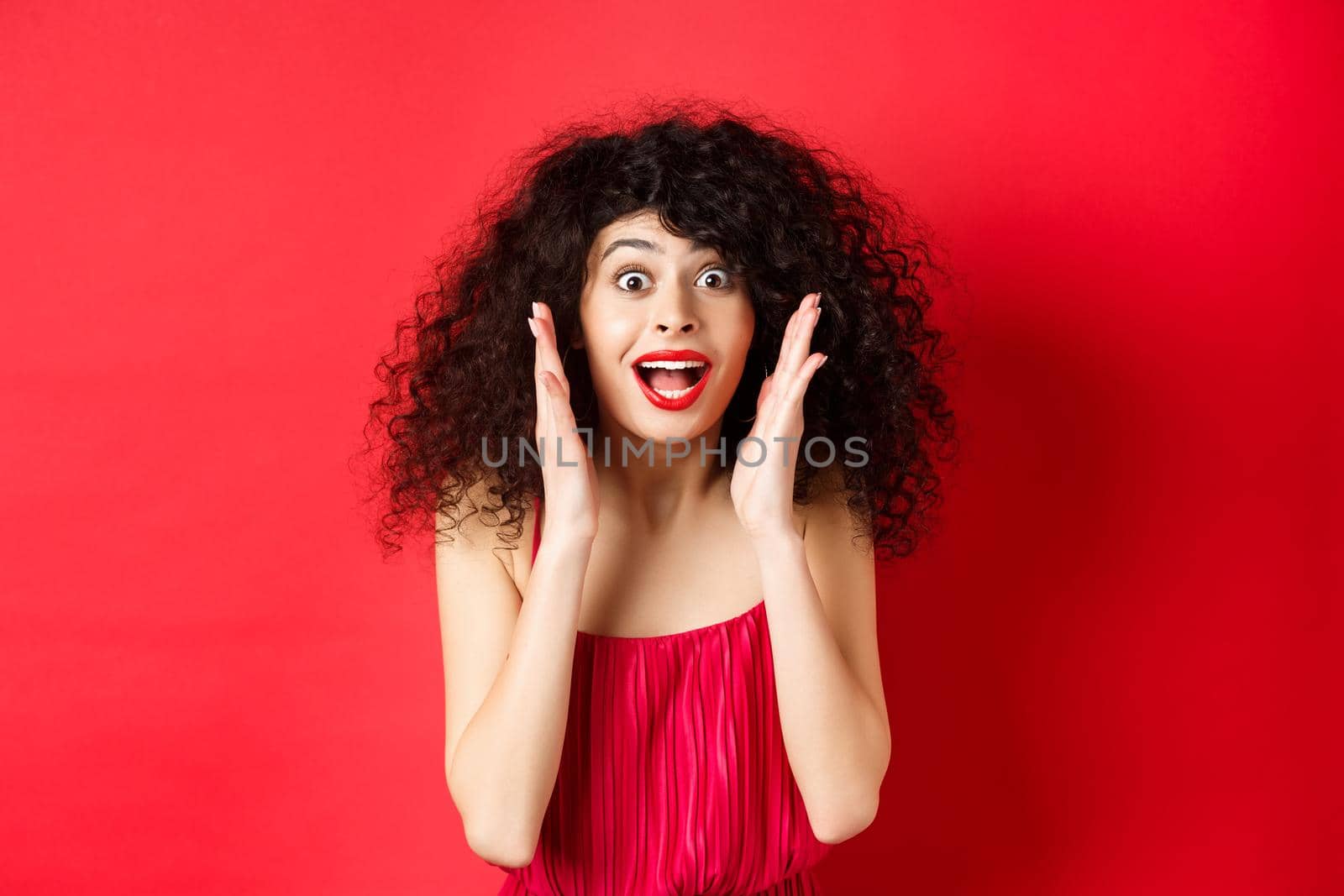 Close-up of surprised beautiful woman, screaming of amazement and looking at promo offer, standing in red dress and lipstick, studio background.