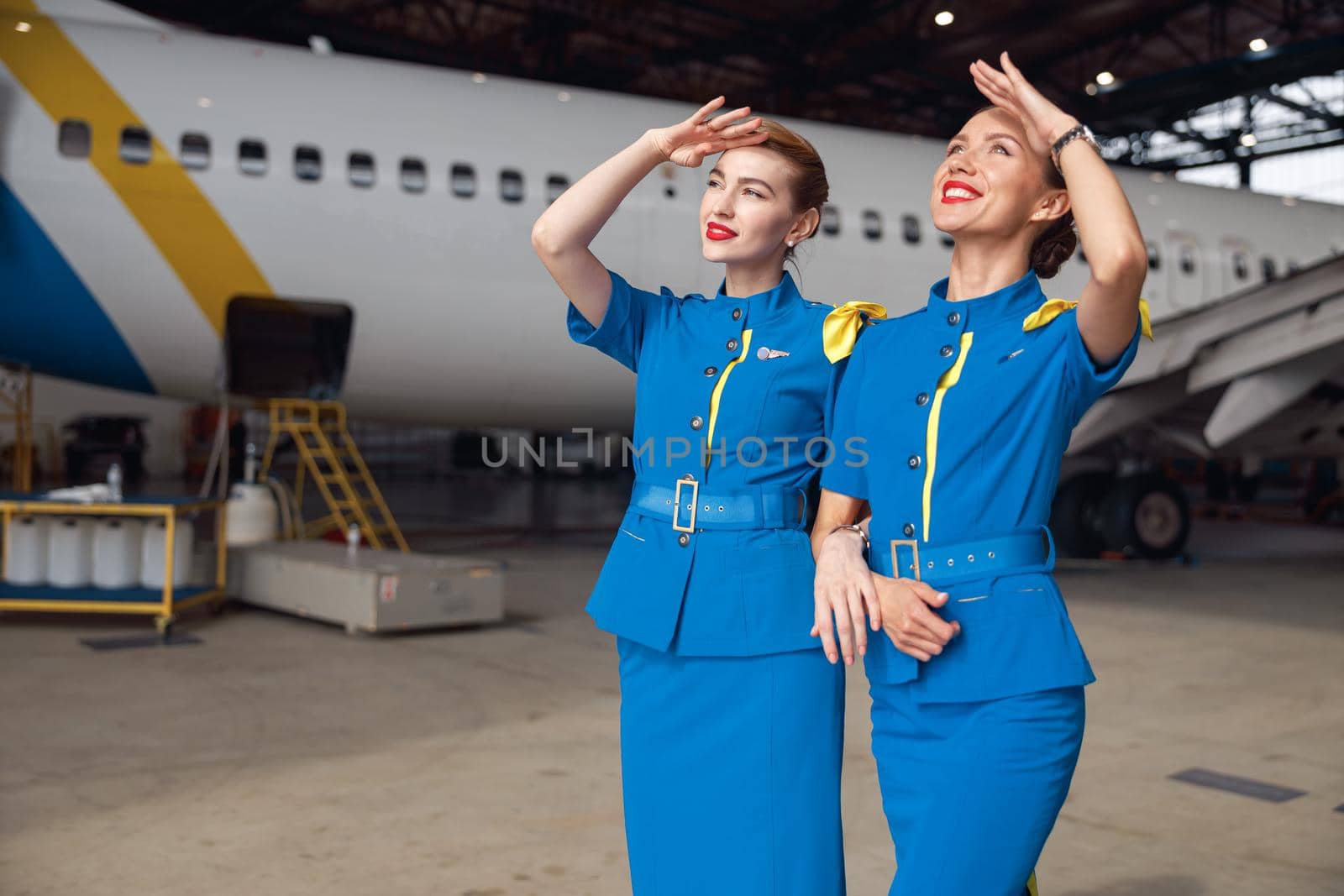 Two air stewardesses in stylish blue uniform smiling while looking up in the sky, standing together in front of passenger aircraft in hangar at the airport. Occupation concept
