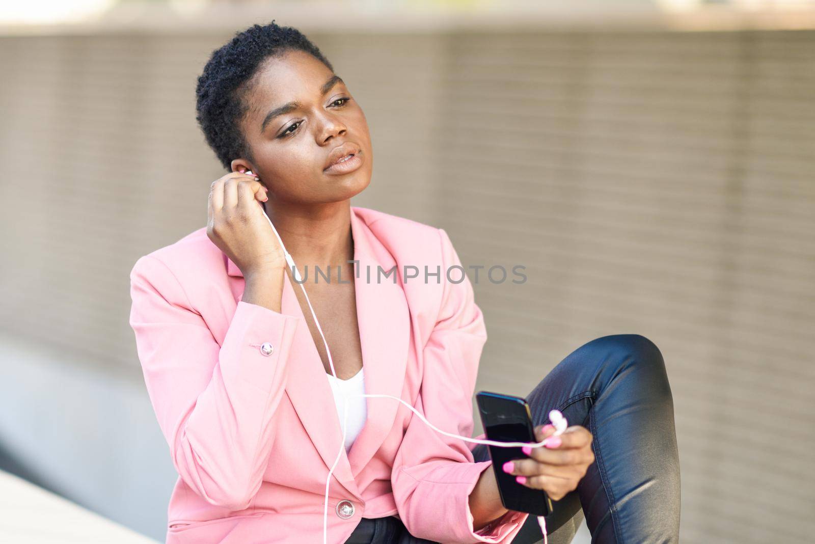 Black businesswoman sitting outdoors using smartphone with earphones. African american female wearing suit with pink jacket.