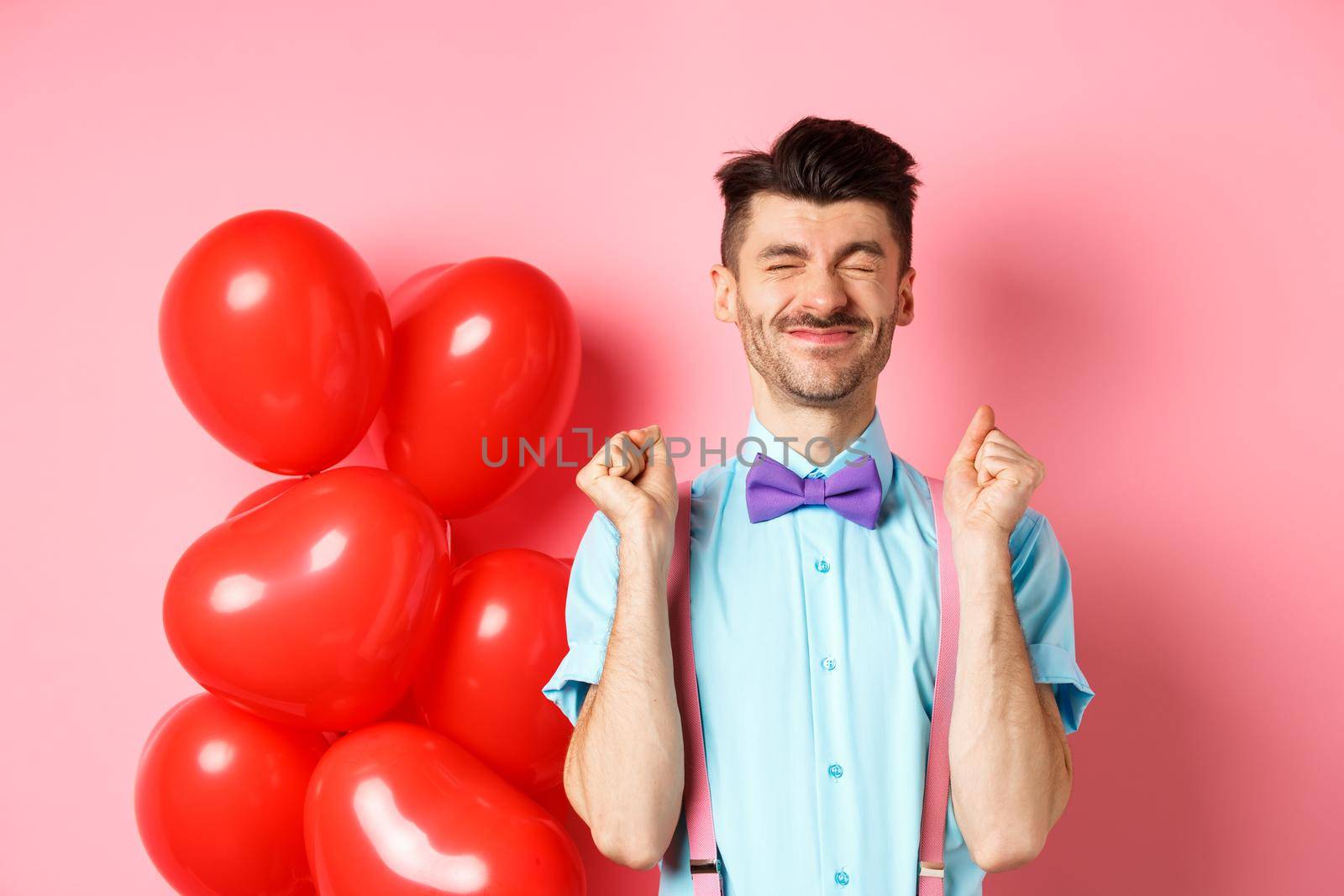 Valentines day concept. Cheerful guy jumping from excitement before romantic date, standing near big red hearts balloons and celebrating, pink background.