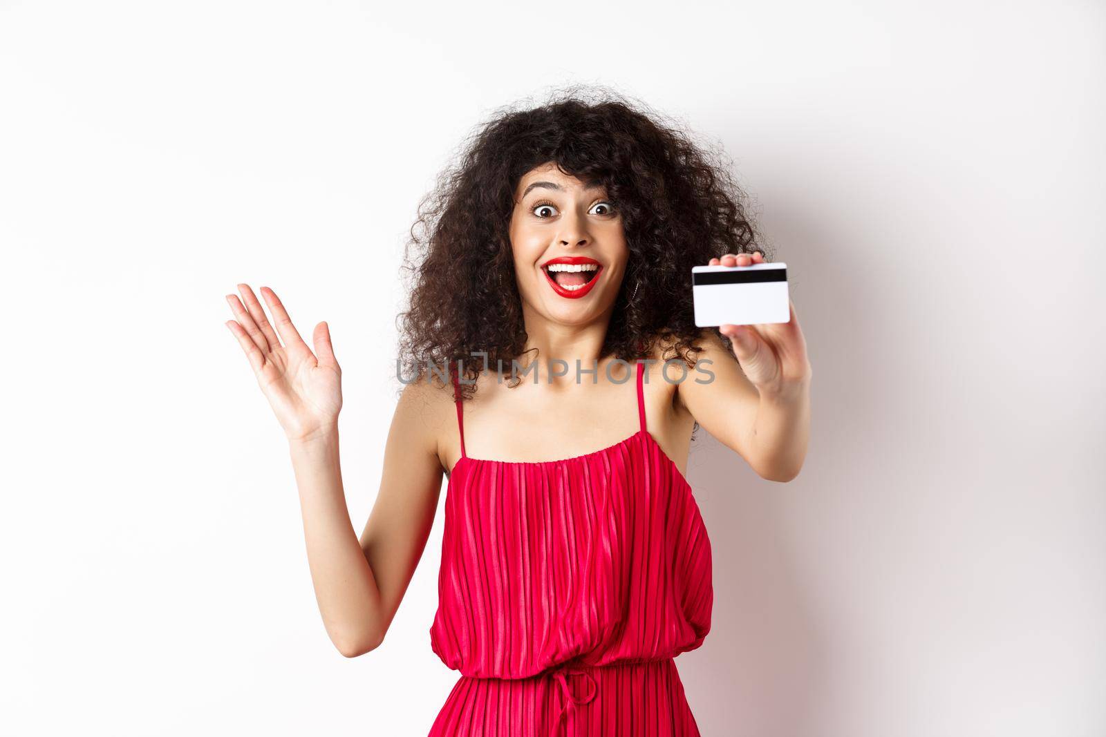 Shopping. Amazed curly-haired woman in red dress, showing plastic credit card and scream from excitement, standing on white background.