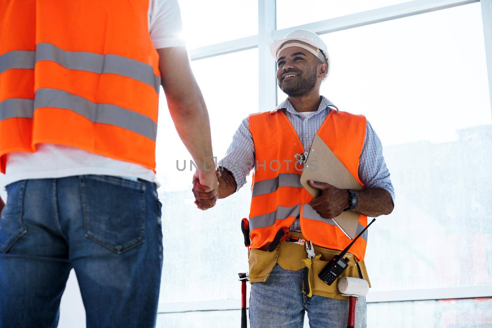 Two men engineers in workwear shaking hands against construction site, close up