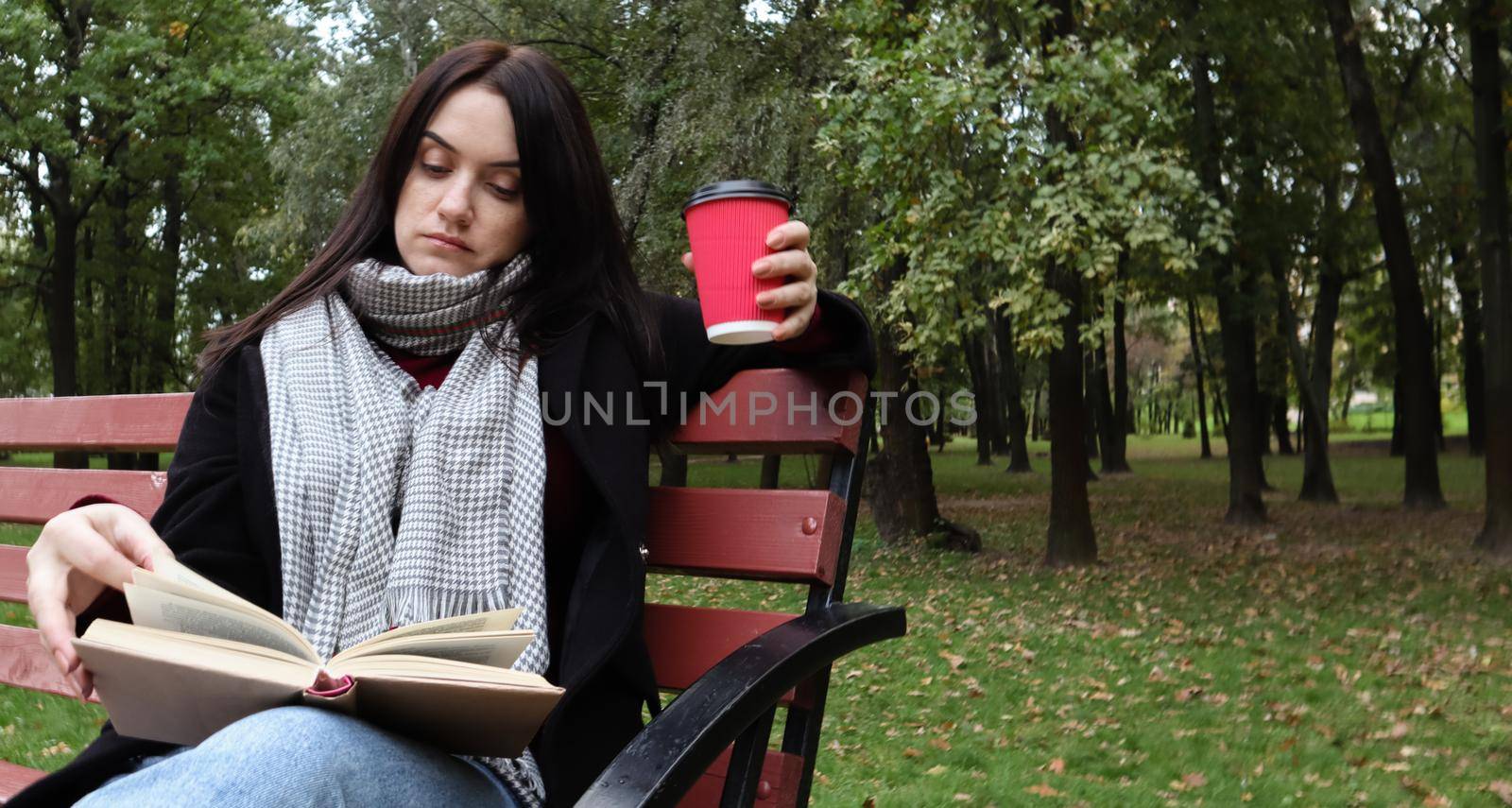 Young woman in jeans, coat and scarf, on a park bench. A woman is reading a book and drinking coffee or other hot drink outdoors alone. by Roshchyn