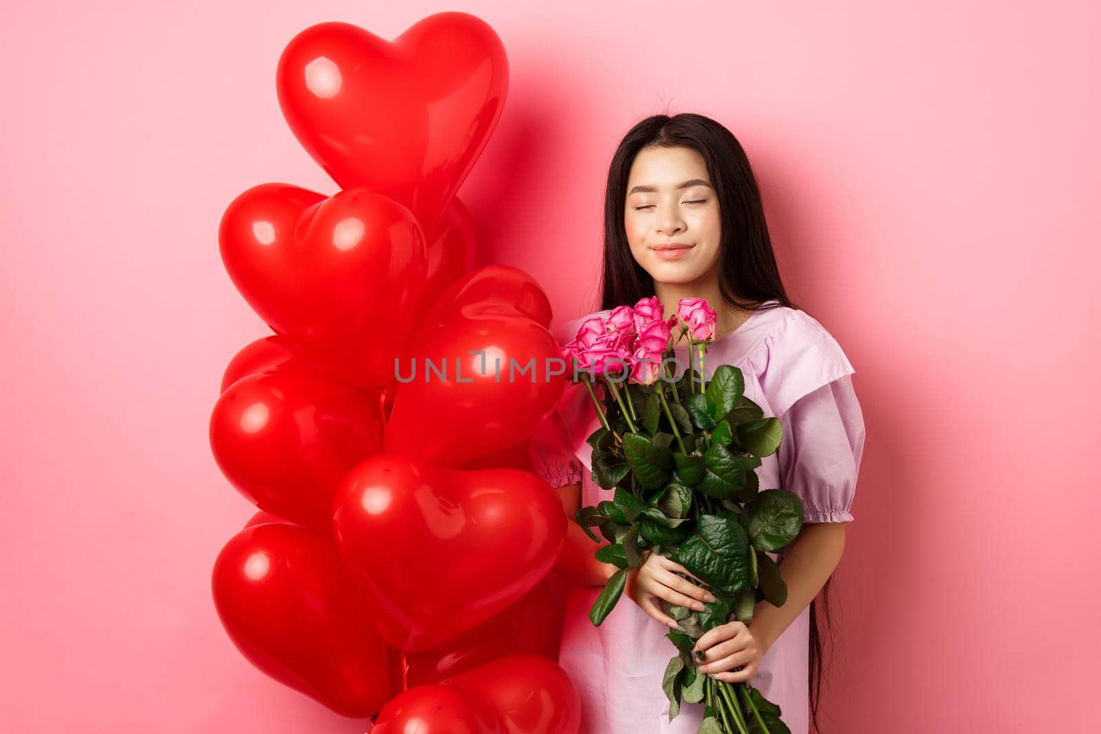 Valentines day concept. Romantic teen asian girl dreaming of love or date, close eyes and smile, holding flowers from lover, receive bouquet of roses and red heart balloons, pink background by Benzoix
