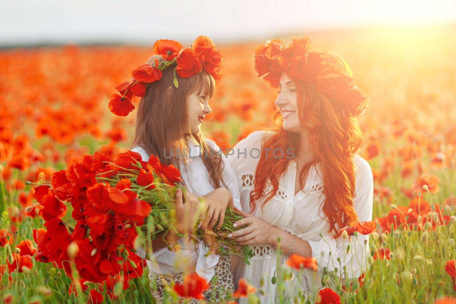 Little girl with redhead mother in white dresses and wreathes poses with bouquet of poppies on poppy field at warm summer sunset