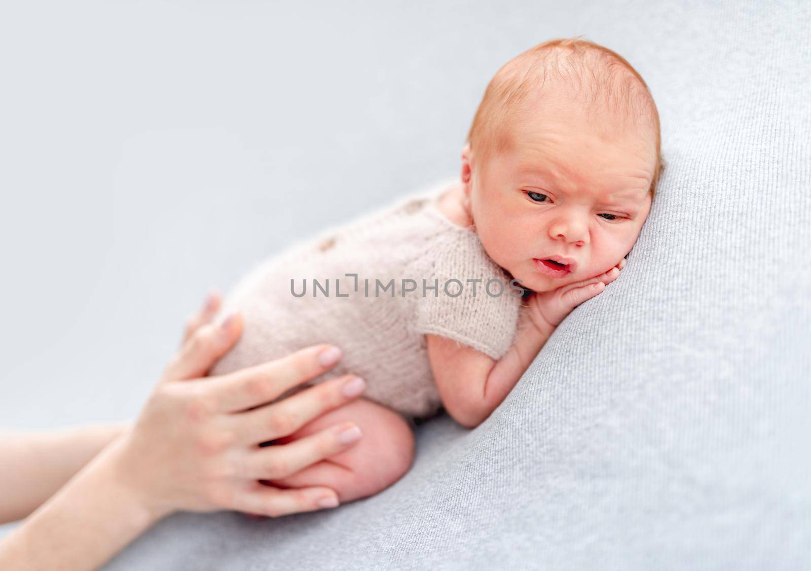 Newborn baby boy wearing knitted beige costume lying on his tummy and his mother hands holding him. Adorable infant child resting and parent cares about him