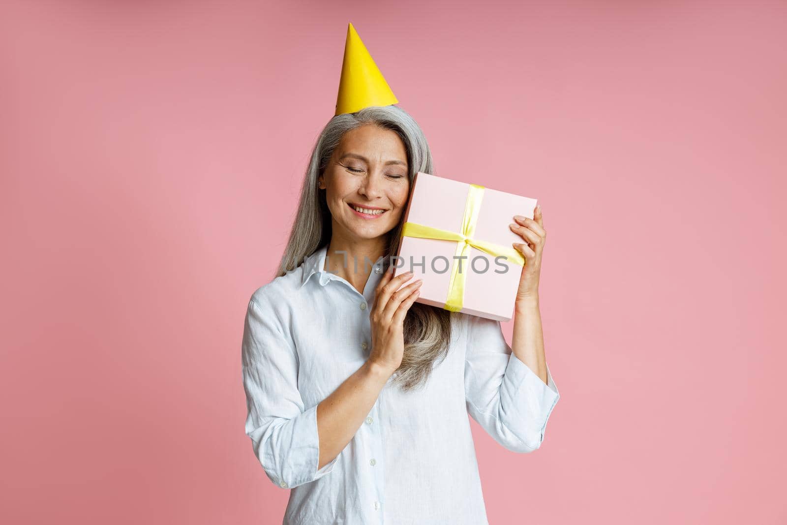 Happy mature Asian lady with long grey hair and yellow party hat holds gift box posing on pink background in studio
