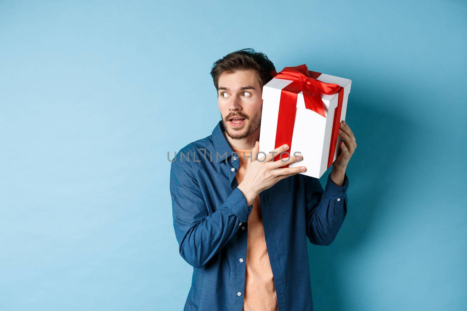 Valentines day. Curious boyfriend holding gift box near ear, trying to guess whats inside surprise present, standing over blue background.