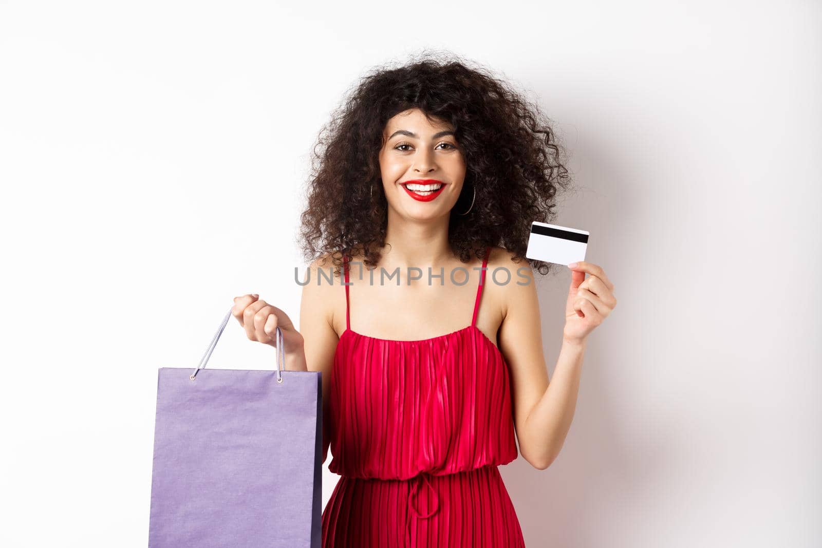 Beautiful female model with curly hair, red dress, showing shopping bag and plastic credit card, white background by Benzoix
